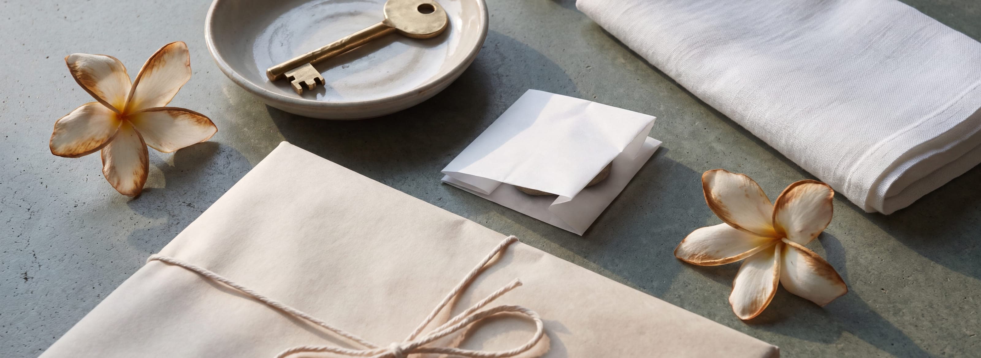 A brass key returned to a ceramic dish beside a wrapped parcel, folded linen and frangipani a day past their best