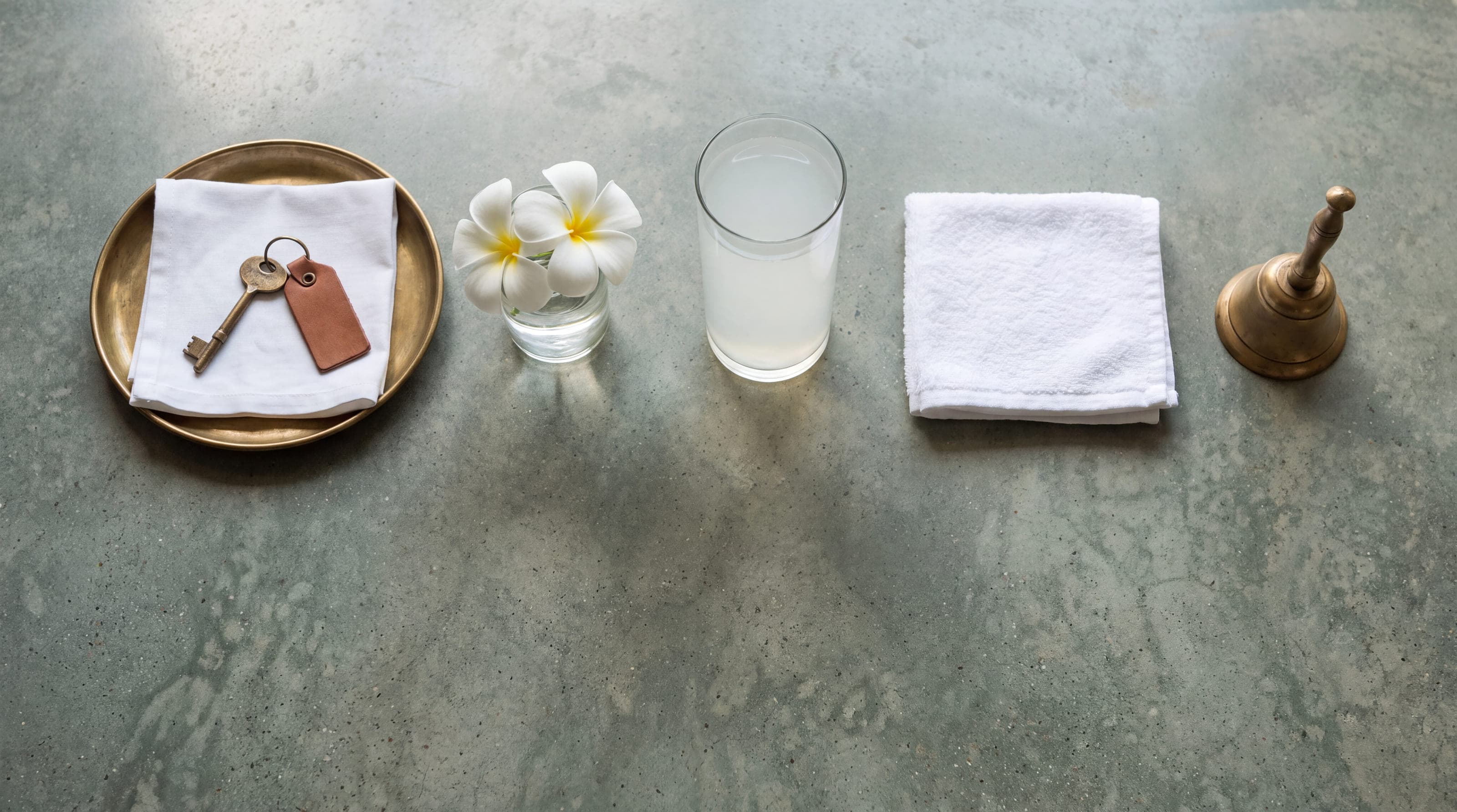 A brass key on a folded cloth on a tray, with frangipani, a glass of king coconut water and a folded towel alongside