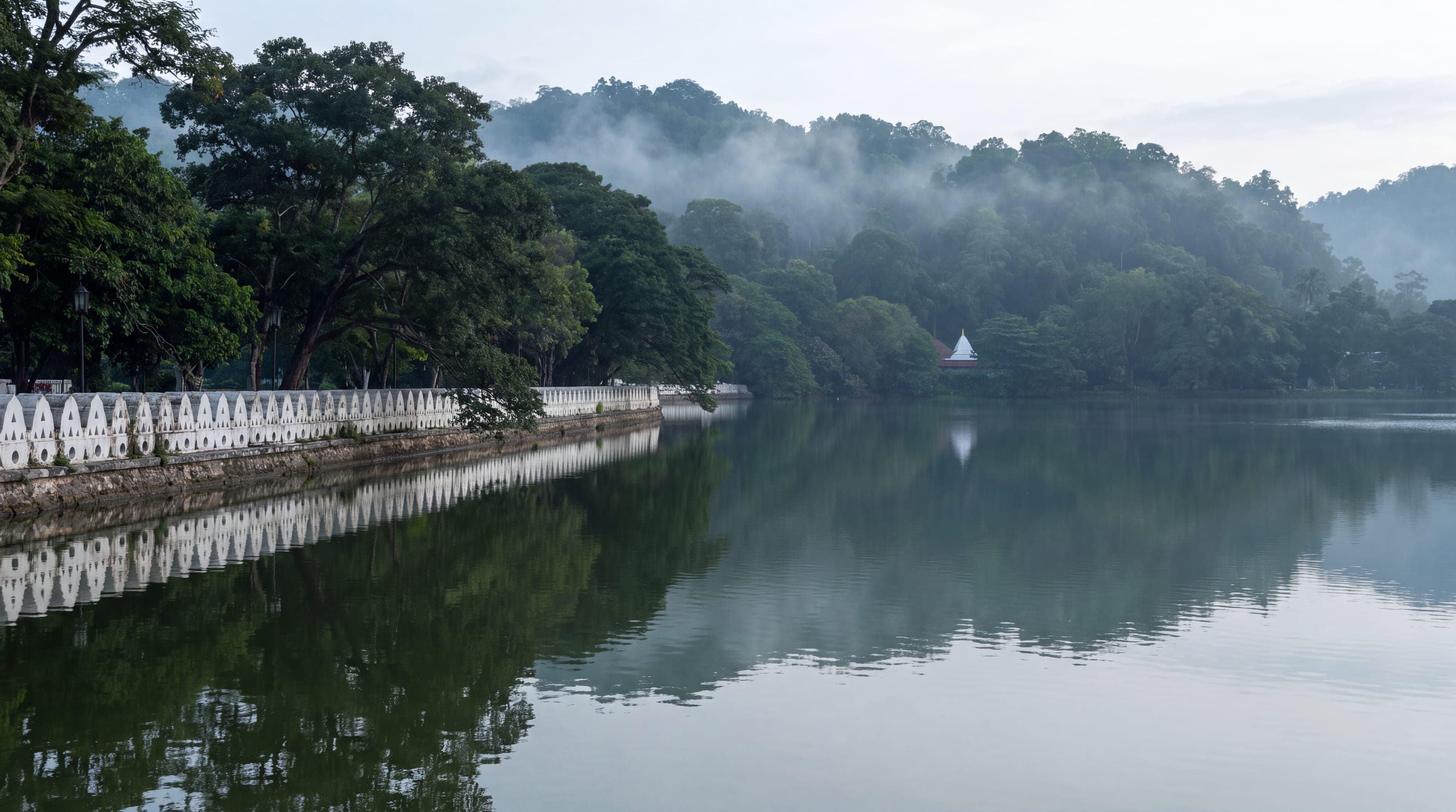 Mist lying on the lake at Kandy at first light, a white balustrade along the near shore and forest rising beyond