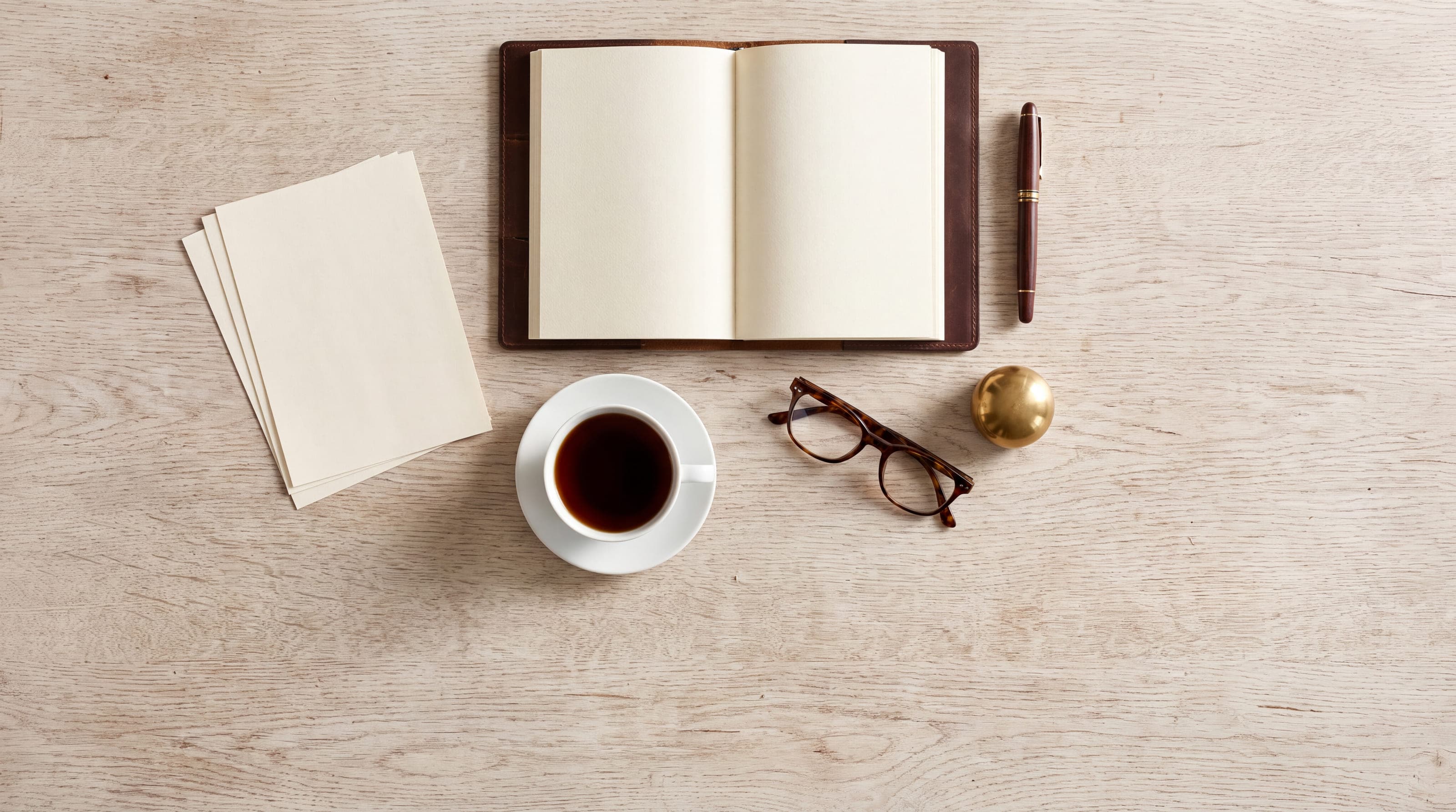 A writing desk from above, an open blank notebook and pen beside loose cards, reading glasses and a cup of coffee